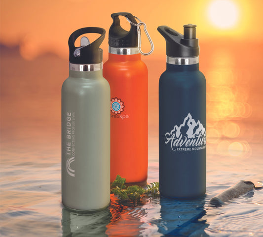 Three branded Nomad vacuum bottles in green, orange, and navy displayed on water at sunset, showcasing custom logos and powder-coated finishes.